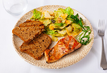 Salmon fillet with lettuce salad served with toasts on plate, over white background.