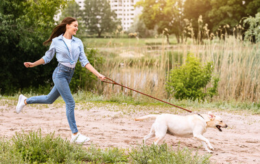 Active Pet Concept. Laughing girl chasing their running dog in park near lake, copy space, full...
