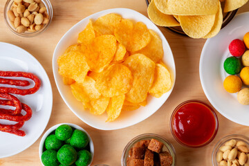 Different snacks such as chips, crackers, salted nuts, wasabi nuts on wooden desk on beige background.