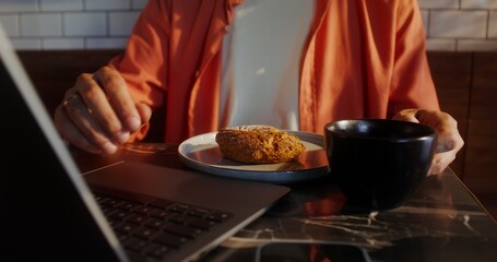 A stylish young man with glasses uses a laptop and eats a donut sitting at a table in a cafe, close-up of a man's hands, no face