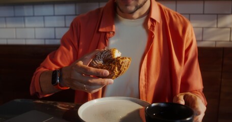 A stylish young man eats a donut and drinks coffee sitting in front of an open laptop in a cafe