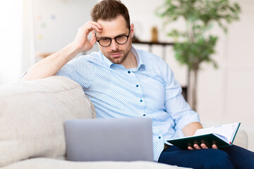 Focused handsome businessman in glasses looking at laptop screen, holding notebook diary, sitting...