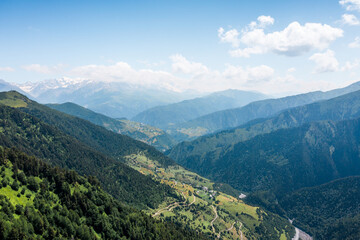 Aerial view of lush green mountains with small villages scattered across valleys. Winding roads connect settlements, creating a picturesque rural landscape under a clear blue sky.