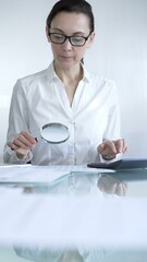 Businesswoman or accountant, wearing white casual shirt and glasses, is using magnifying glass and calculator to analyze financial documents in office setting. Audit and taxes in business