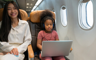 African girl wearing headphones watching laptop screen on airplane while Asian woman beside her smiling. Relaxation and entertainment during airline travel journey concept