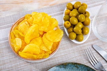There is portion of yellow crispy potato chips on small plate. Popular snack is complemented by bowl with delicate green olives