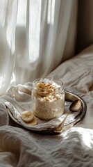 Creamy oatmeal in a glass jar with banana and coconut served on a glossy metall tray, on linen bedding, slow living and lazy breakfast atmosphere in pastel tones
