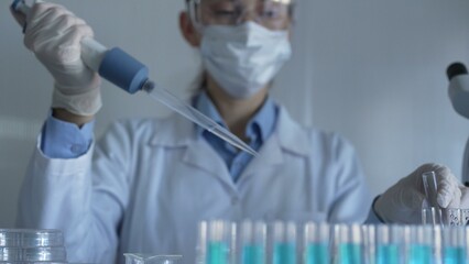 Scientist woman, wearing a lab coat, mask, blue gloves and safety glasses, is using a micropipette...