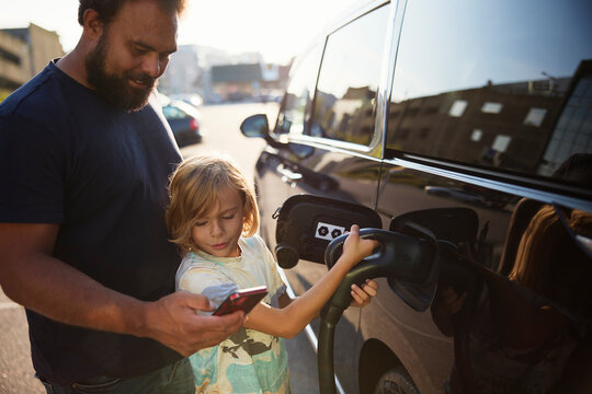 Father showing smart phone to son charging electric car with plug at street
