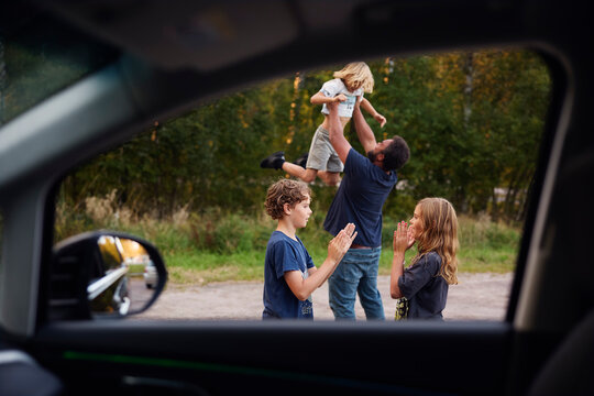 Happy brother and sister playing clapping game while father lifting son seen through car window