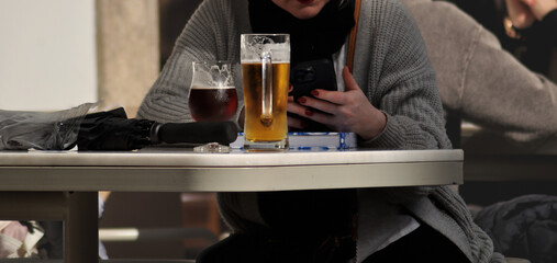 Person sitting at a terrace table with a cell phone in hand, a glass and a mug of beer on the table, umbrella also resting on the table