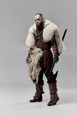 Fototapeta premium Full-length portrait of determined man, medieval Viking dressed in leather armor and fur cloak, standing with axe against grey studio background. Concept of historical portrait, Nordic mythology