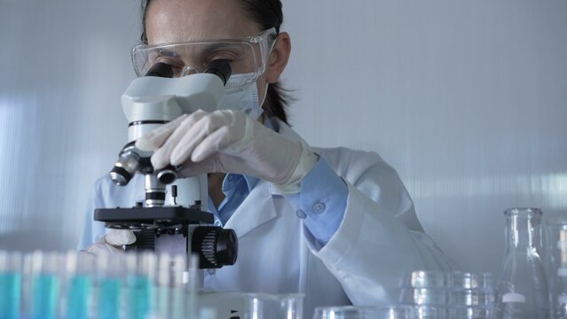 Scientist woman, wearing a lab coat, mask, white gloves and safety glasses, is using a microscope surrounded by test tubes in laboratory. Portrait view. Medicine and science concept