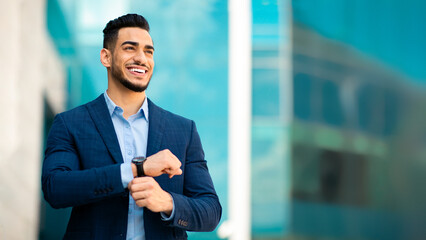 Successful arabic entrepreneur young handsome man in suit with expensive watch on his wrist looking at copy space and smiling, walking on the street next to business building, panorama