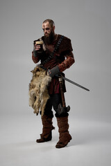 Viking warrior standing with sword and axe, holding glass filled with beer, dressed in fur cloak and leather armor against studio background. Concept of Nordic traditions, medieval warriors rest.