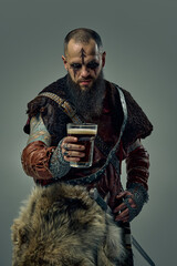 Portrait of medieval Viking warrior dressed in fur cloak and leather armor, standing with sword, holding glass of beer, against studio background. Concept of Nordic traditions, beer festival
