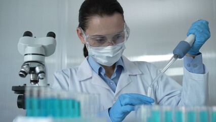 Scientist woman wearing a lab coat, mask, and safety glasses is using a micropipette to drop a blue...