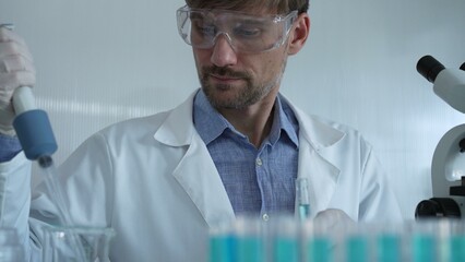 Scientist man wearing a lab coat and safety glasses is using a micropipette to drop a liquid into a...