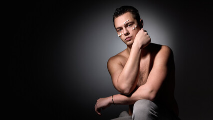 Naklejka premium Shirtless young man sitting with arm resting on knee, white pills on face, looking into camera with moody lighting against dark studio background. Concept of selfcare, natural beauty, cosmetology.