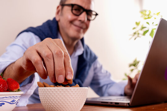 Happy senior man working on laptop and eating a healthy almond snack at his desk. Concept of active retirement, brain food, and corporate wellness.