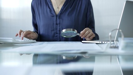Businesswoman analyzing financial data with magnifying glass and calculator, performing accounting...