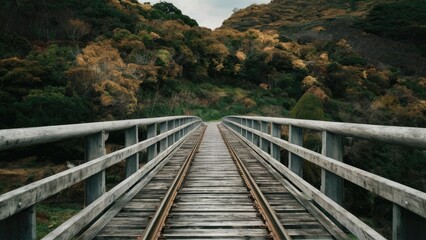 Fototapeta premium Wooden bridge with small rails extending across it surrounded by lush green and autumn foliage in a scenic landscape under a cloudy sky.