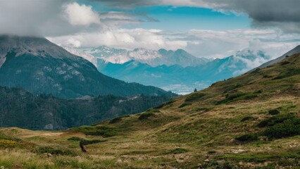 Fototapeta premium Lush green mountain vegetation with dramatic clouds overhead contrasting against a serene blue sky and distant peaks in the background