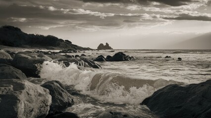 Obraz premium Monochrome coastal scene featuring dark rocks and white frothy waves in the foreground, with distant formations under a cloudy sky.