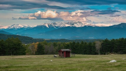 Scenic mountain landscape with snow-capped peaks in the background green field and rustic red cabin under a dramatic sky with clouds