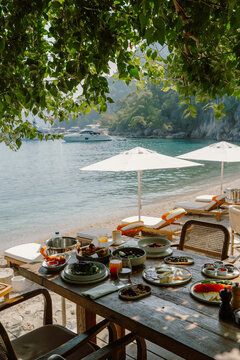 A picturesque seaside breakfast setup in Fethiye, T&uuml;rkiye, featuring fresh fruits, cheeses, bread, and olives on a rustic wooden table with ocean views, white umbrellas, and yachts in the background