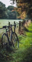 Fototapeta premium Black Road Bike Leaning Against Fence in Green Grassy Countryside with Trees and Soft Focus Background