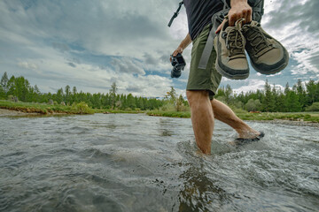 A tourist traveler crosses the river holding his shoes in his hands, against the background of the sky, forest and mountains. A man crosses a stream barefoot in the forest.
