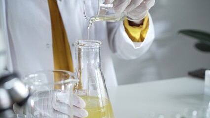 A scientist wearing a lab coat and white gloves is pouring a yellow oily liquid from one beaker to another in a laboratory setting, close up. Science and medicine