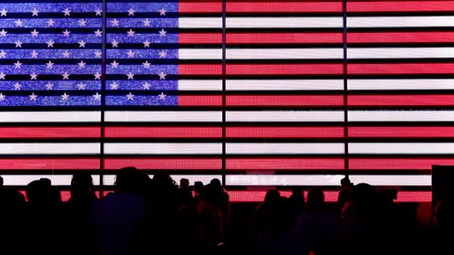 A lively crowd gathers in front of a massive illuminated American flag display at night, showcasing patriotism, urban energy, and the vibrant atmosphere of a bustling city