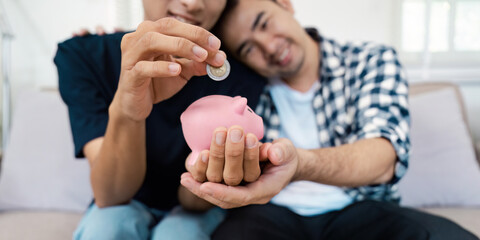 Diverse LGBTQIA couple saving money together, placing coins into a pink piggy bank at home.