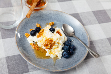Fresh farm cottage cheese filled with large amount of liquid summer honey is served on plate, decorated with walnut and blueberry