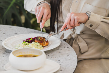 A woman is elegantly cutting into a beautifully plated meal, featuring salmon, mashed potatoes, and soup, all set on a stylish table.