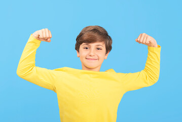 Boy raising arms with clenched fists, showing biceps, smiling face, yellow t-shirt, light blue background, winner gesture