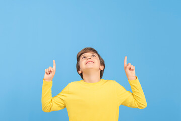 Charming young boy pointing and pointing with arms and fingers upwards in yellow sweater, showing something interesting and surprising, isolated on blue background
