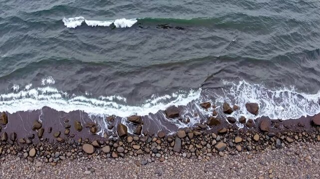 Bird's eye view, moves parallel to the shore, capturing rolling waves tumbling over scattered stones, the neutral palette blending sky, sea, and earth.

