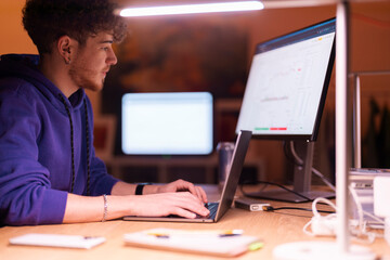 A young man in a purple hoodie sits at a wooden desk in a dimly lit office, working on a laptop while analyzing financial graphs displayed on a large screen.