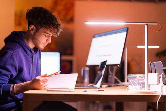 A young man in a purple hoodie sits at a desk in a warmly lit office, focused on his laptop while holding a pencil, with a monitor displaying financial charts nearby.