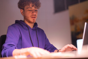 A young man in a purple hoodie sits at a wooden desk in a dimly lit office, intently looking at a large monitor while working on a project.