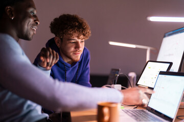 Two young professionals sit at a shared desk in a dimly lit office, reviewing stock market data displayed on multiple screens while discussing investment strategies.