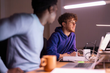 Two young professionals sit at a shared desk in a dimly lit office, reviewing stock market data displayed on multiple screens while discussing investment strategies.