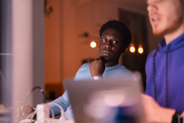 Two young men work together at a desk in a modern office, reviewing data on multiple screens while collaborating on a financial or business-related project.