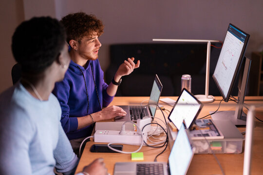 Two young men sit at a desk in a dimly lit office, discussing financial data displayed on multiple screens while analyzing stock market trends and investment opportunities. - Powered by Adobe