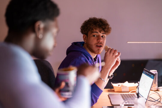 Two young professionals sit at their desks in a modern office, eating and discussing while working on their laptops, with multiple monitors displaying stock data.