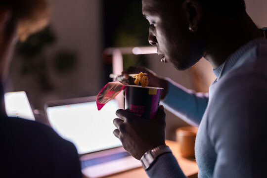 A man in a blue sweater is eating instant noodles from a cup using a fork while working on a laptop at night in a dimly lit room with a warm ambiance.