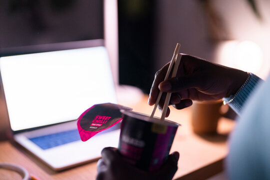 A close-up of a hand holding chopsticks and picking up noodles from a cup of sweet chili instant noodles beside an open laptop in a dimly lit workspace.